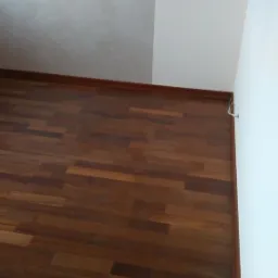 Ukończona podłoga z ciemnego, lakierowanego parkietu ułożonego w cegiełkę, widoczna przy ścianie z dekoracyjnym tynkiem i białym narożnikiem. Listwa przypodłogowa w kolorze parkietu.