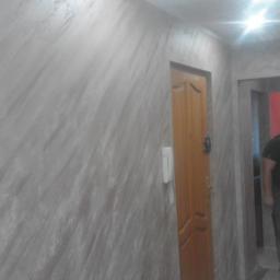 Rafał Bulo - Ściana w korytarzu wykończona dekoracyjnym tynkiem w odcieniach szarości, z widoczną drewnianą ościeżnicą drzwi i domofonem na ścianie.