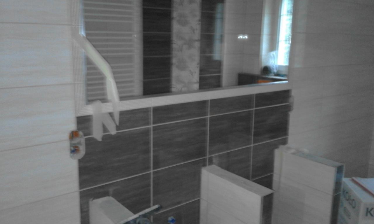 Fragment łazienki w trakcie wykańczania, widoczne płytki ścienne w odcieniach szarości i beżu, lustro z odbiciem okna, oraz karton z napisem 'Koło' sugerujący elementy wyposażenia łazienki.