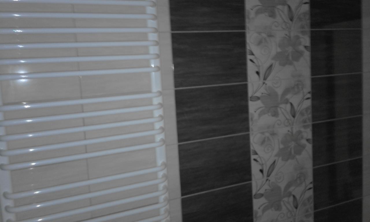 Biały grzejnik drabinkowy łazienkowy na tle ściany z płytek w odcieniach beżu, szarości i wzoru kwiatowego.