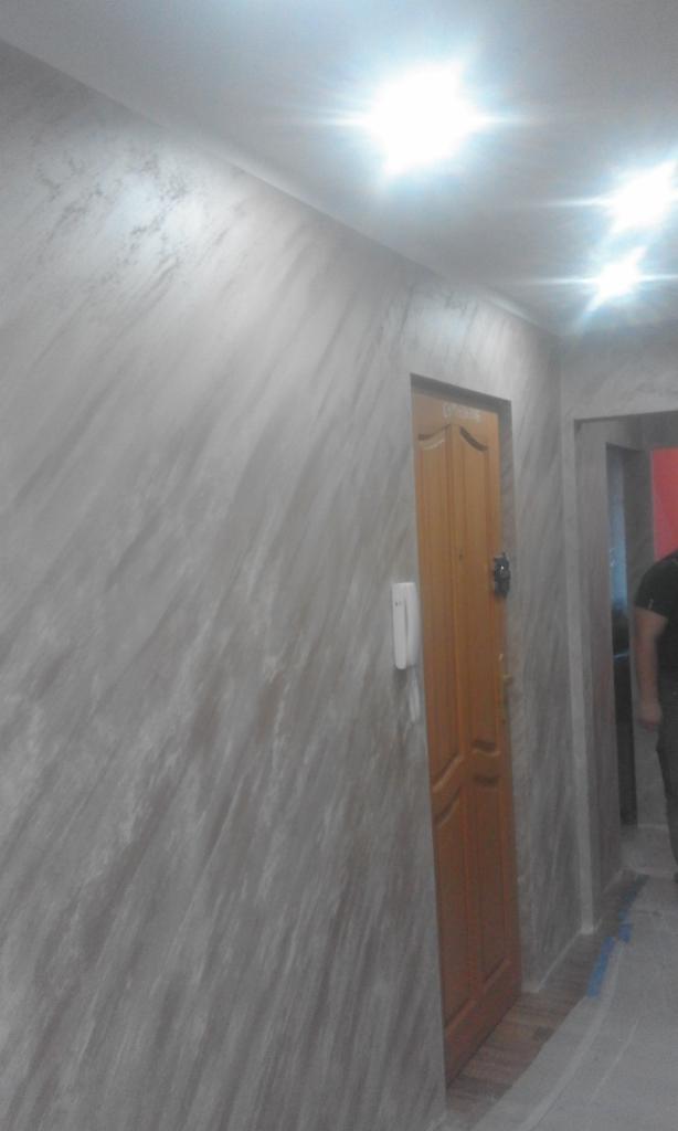 Ściana w korytarzu wykończona dekoracyjnym tynkiem w odcieniach szarości, z widoczną drewnianą ościeżnicą drzwi i domofonem na ścianie.