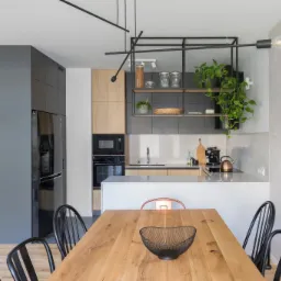 Nowoczesny salon z kuchnią i antresolą: drewniany stół z czarnymi krzesłami, minimalistyczna kuchnia z drewnianymi i szarymi szafkami, czarna lodówka, oryginalne oświetlenie sufitowe, betonowa...