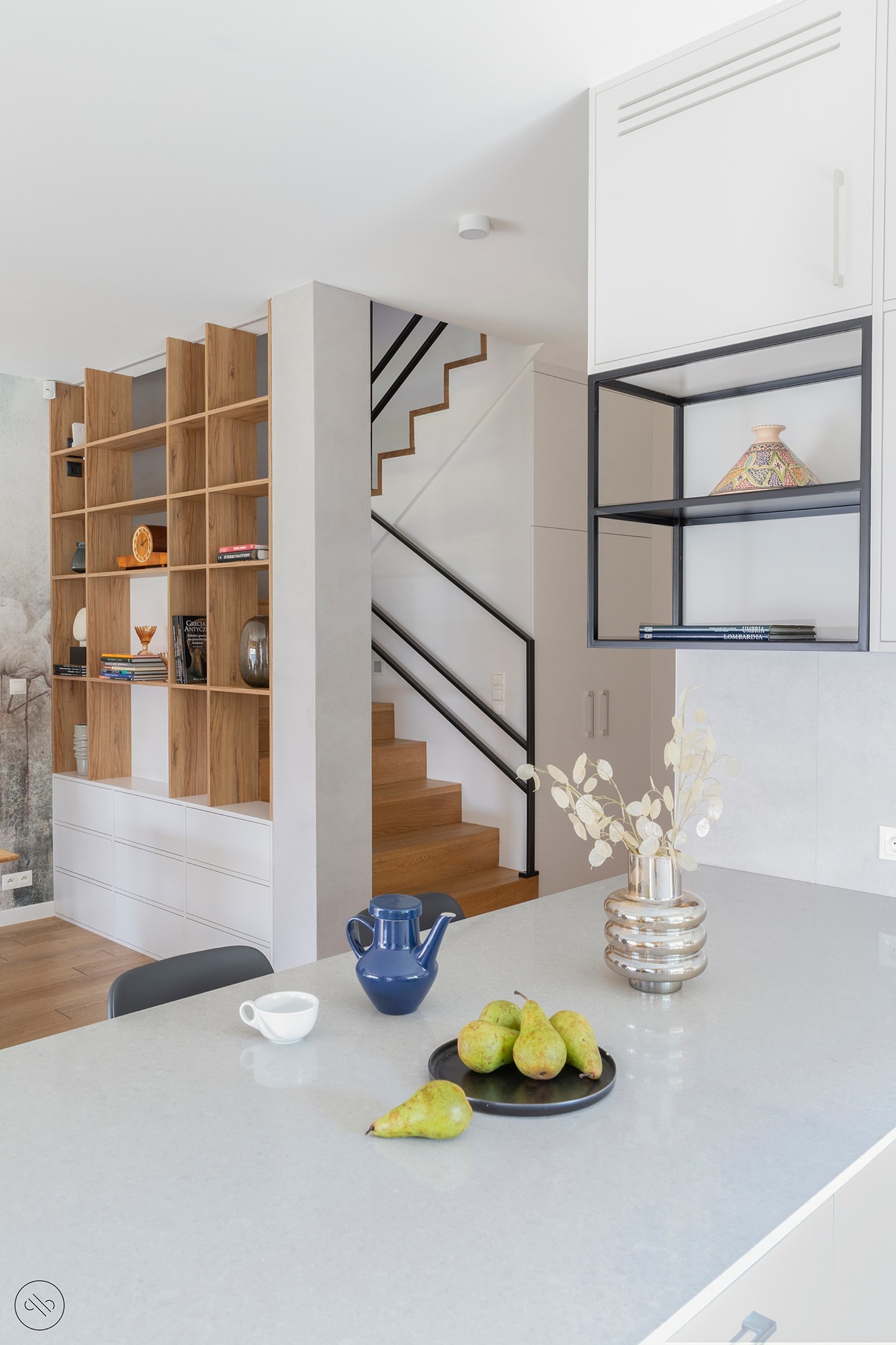 Jasne wnętrze salonu z kuchnią w domu w Warszawie: drewniany regał z książkami i dekoracjami, schody z czarną balustradą, na blacie dzbanek, filiżanka, gruszki i srebrny wazon z suszonymi kwiatami.