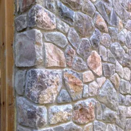 Ściana wykończona nieregularnymi kamieniami w odcieniach szarości, beżu i brązu, z widoczną drewnianą ramą po lewej stronie.