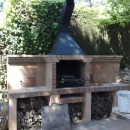 Murowany grill ogrodowy z cegły, z czarnym kominem, drewnem na opał w dolnej części i zielonym żywopłotem w tle.