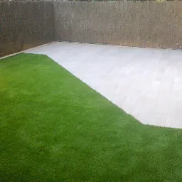 Wyłożony jasnymi płytami taras z fragmentem sztucznej trawy, ogrodzony matą trzcinową i kamiennym murkiem.