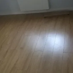 Ułożone panele podłogowe w jasnym odcieniu drewna, widoczne listwy przypodłogowe i fragment niezamontowanych paneli przy ścianie.