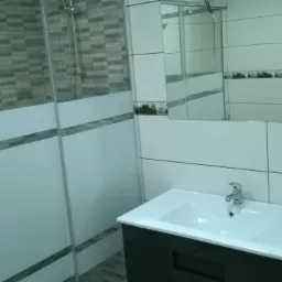 Nowoczesna łazienka z kabiną prysznicową, umywalką na szafce i białymi płytkami z dekoracyjnym pasem, w lustrze odbicie mężczyzny.