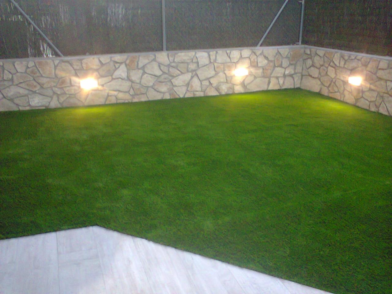 Wykończony ogródek z zieloną sztuczną trawą, oświetlonym murem z kamienia i fragmentem jasnej podłogi z desek.