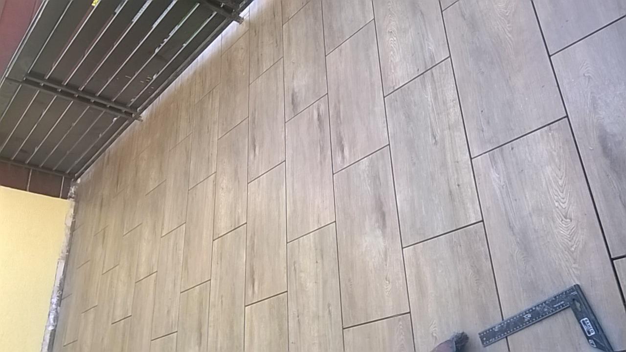 Ściana wyłożona podłużnymi, szarymi płytkami imitującymi drewno, z widocznym kątownikiem i fragmentem rusztowania, ukazująca etap prac wykończeniowych.