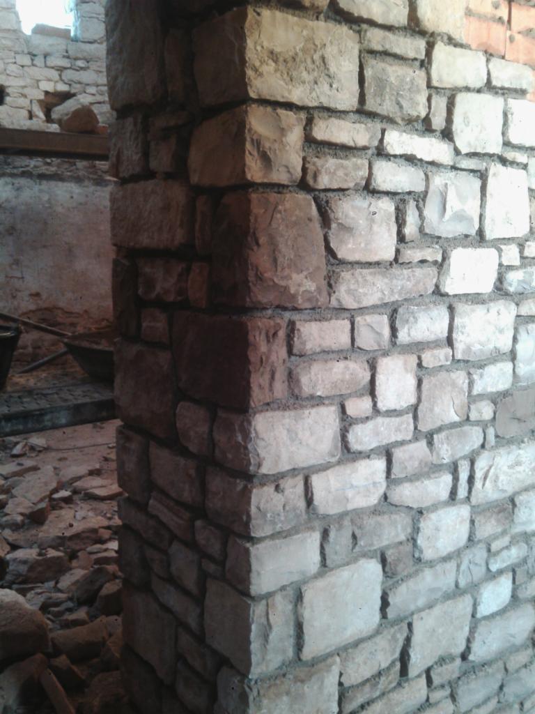 Fragment narożnika ściany wykończonej nieregularnym kamieniem w odcieniach szarości i beżu, z widocznymi spoinami, w tle widoczny gruz i elementy konstrukcyjne budynku w trakcie prac.