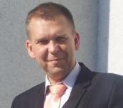 Portret mężczyzny w garniturze z krawatem w odcieniach brzoskwini, z krótkimi włosami i lekkim uśmiechem, na tle jasnej ściany. Ujęcie od ramion w górę.