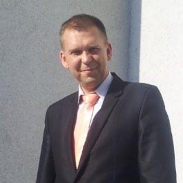 Portret mężczyzny w czarnym garniturze i jasnym krawacie na tle jasnej ściany. Uśmiechnięty, z krótkimi włosami.