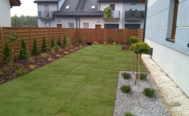 Świeżo założony trawnik z rolki przed domem, obrzeża wykończone korą i kamieniami, drewniane ogrodzenie w tle.