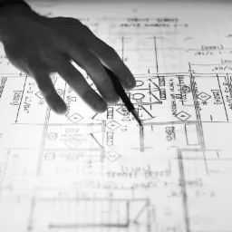 Ręka z ołówkiem wskazuje na plan architektoniczny z detalami łazienki i oznaczeniami, leżący na podświetlonym stole kreślarskim. Czarno-biała tonacja.