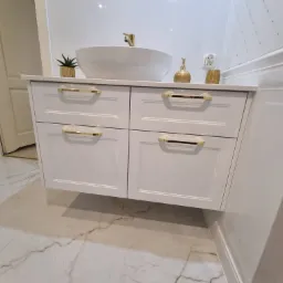 Biała szafka łazienkowa z czterema szufladami i złotymi uchwytami pod umywalką w kształcie misy, na marmurowej podłodze, z widocznymi elementami dekoracyjnymi.