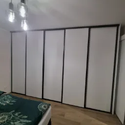 Biała szafa przesuwna z czarnymi obramowaniami, widoczna w sypialni z pościelą w odcieniach zieleni i wzorem łapacza snów, oświetlona przez nowoczesną lampę sufitową.