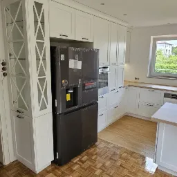 Nowoczesna biała kuchnia z zabudową meblową, czarną lodówką w folii ochronnej i parkietem w jodełkę.