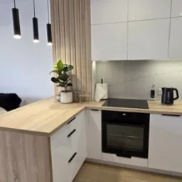 Nowoczesna kuchnia z białymi szafkami i drewnianym blatem, oświetlona wiszącymi lampami. Elementy dekoracyjne: roślina doniczkowa i drewniana ścianka działowa.