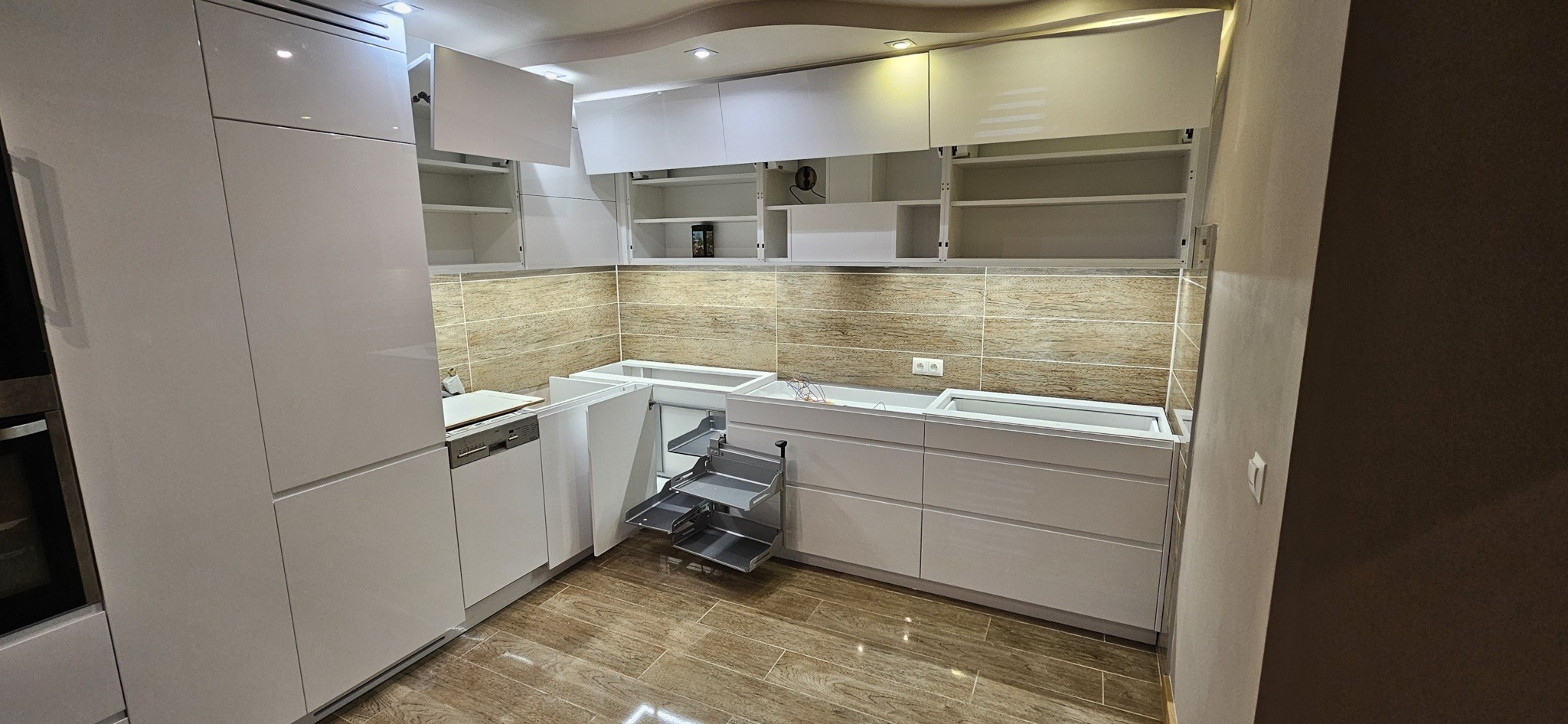 Wnętrze kuchni w trakcie montażu, z otwartymi szafkami górnymi i dolnymi, widoczny system wysuwanych półek narożnych, jasne, błyszczące fronty, płytki na ścianie imitujące drewno.