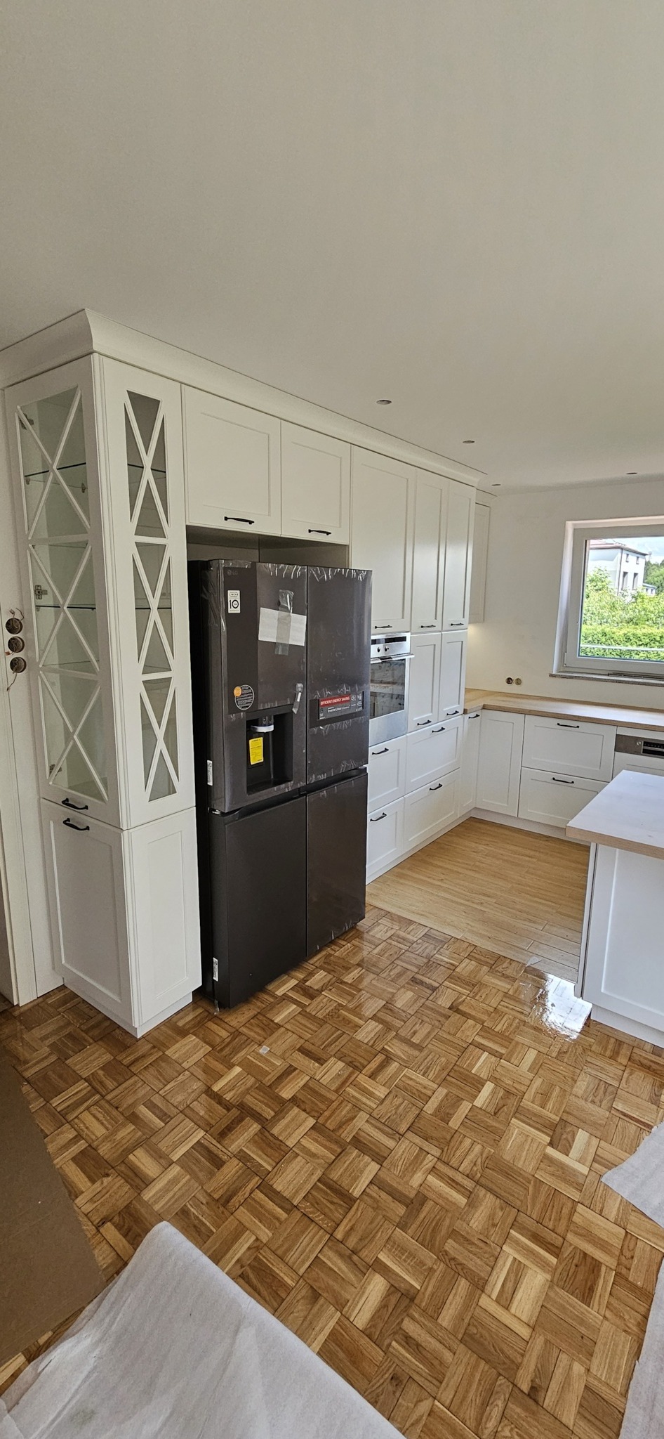 Nowoczesna biała kuchnia z zabudową meblową, czarną lodówką w folii ochronnej i parkietem w jodełkę.