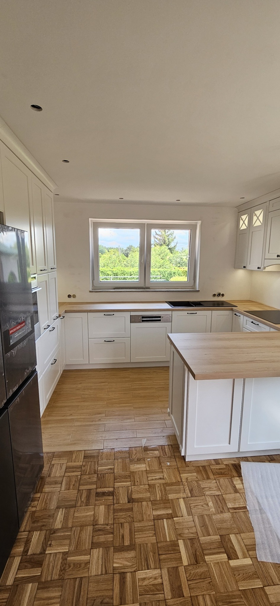 Jasna kuchnia z białymi szafkami i drewnianym blatem, widok na okno z zielenią na zewnątrz, parkiet z ciemnego drewna ułożony w kwadraty, nowoczesna lodówka po lewej.