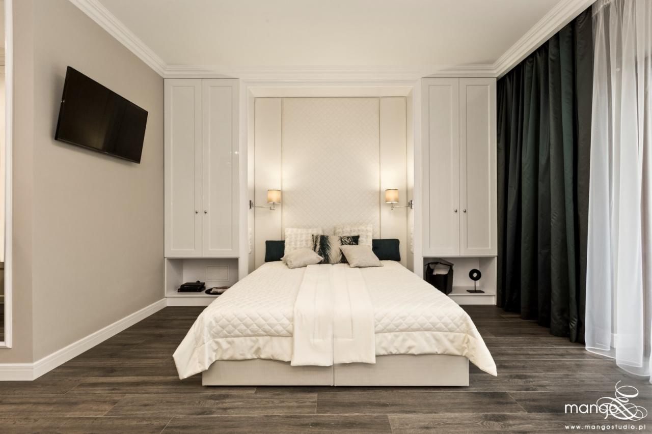 Elegancka sypialnia z dużym łóżkiem, wbudowanymi szafami po bokach, telewizorem na ścianie i ciemnymi zasłonami przy oknie.