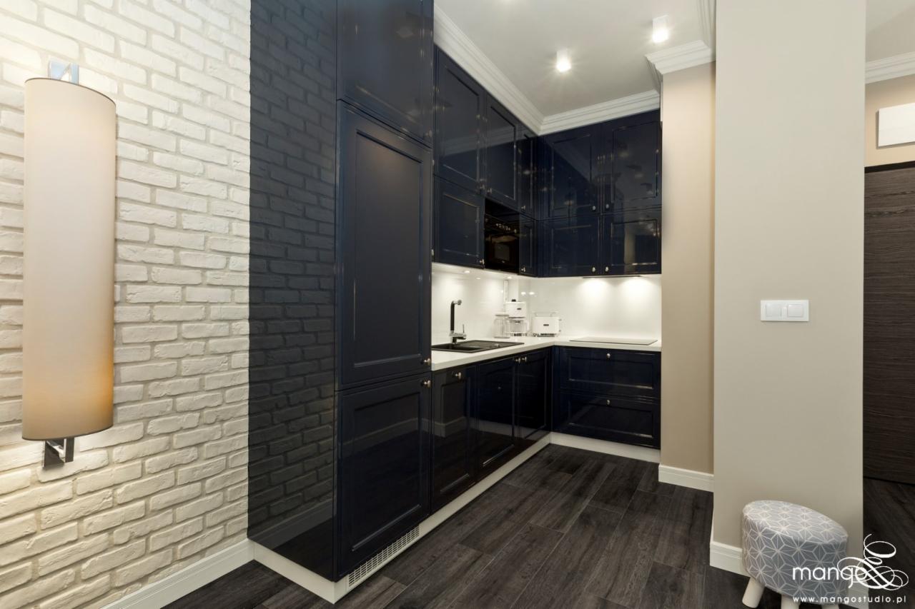 Elegancka, granatowa kuchnia z lakierowanymi frontami w Krakowie, kontrastująca z białą cegłą na ścianie i drewnopodobną podłogą, oświetlona lampą ścienną.