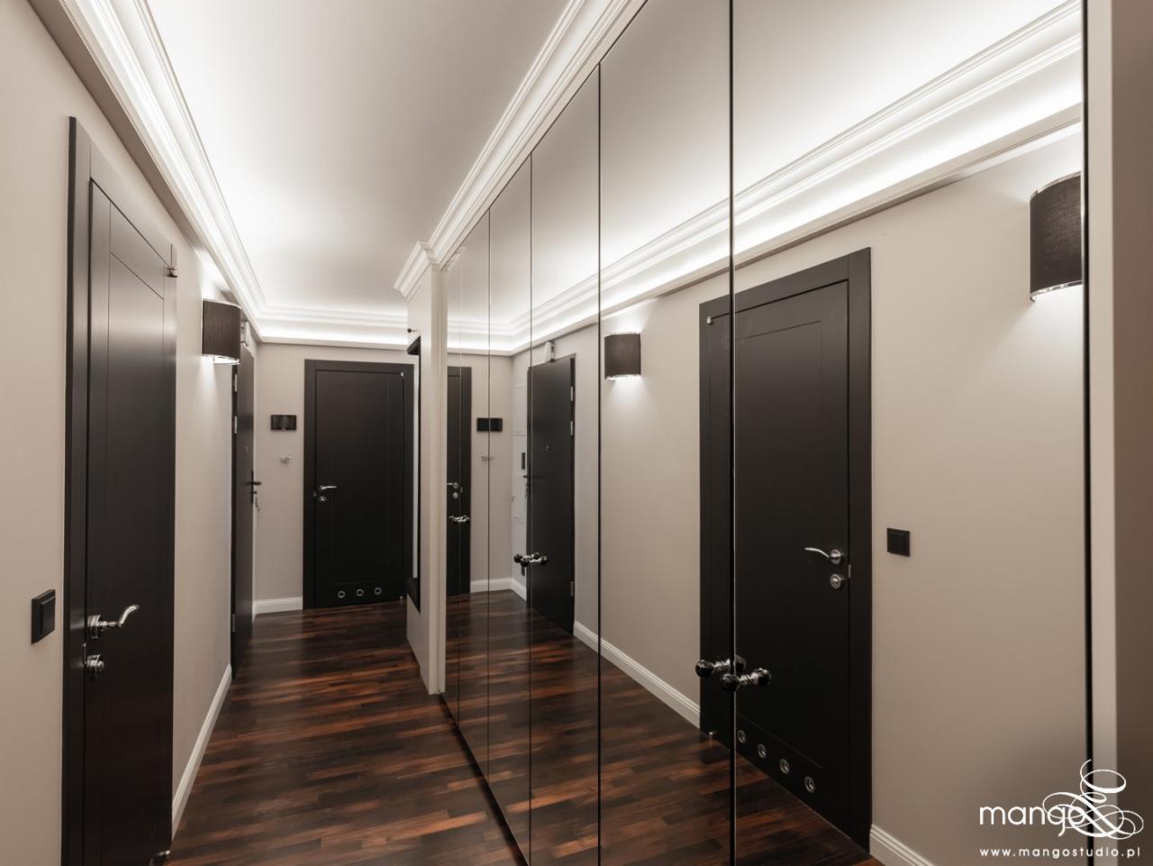 Korytarz z ciemnymi drzwiami, parkietem w jodełkę i lustrzanymi szafami wnękowymi, oświetlony kinkietami i listwami LED.