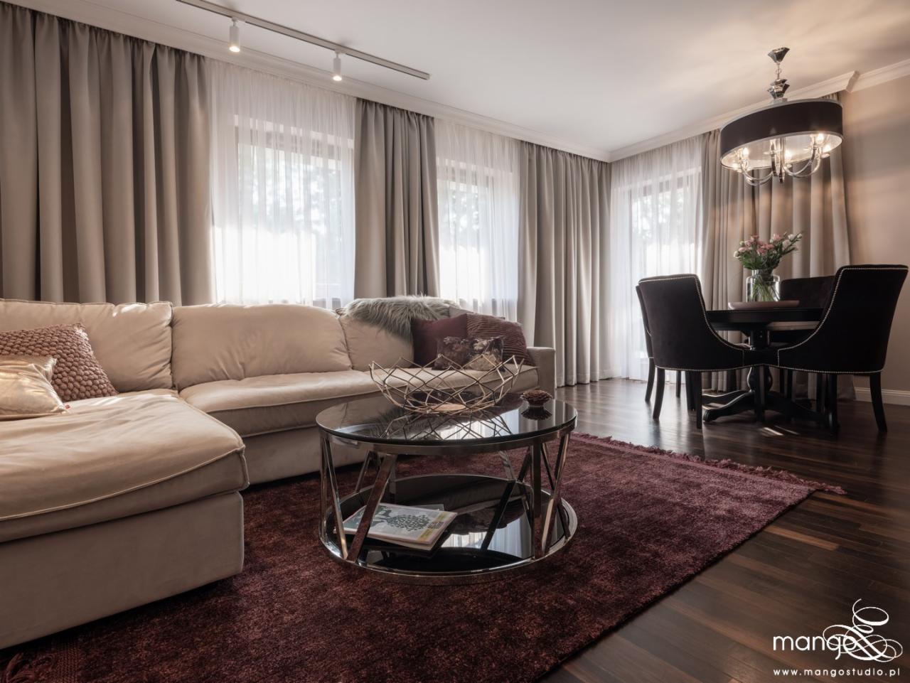 Salon z beżową sofą narożną, okrągłym stolikiem kawowym z metalową konstrukcją i ciemnofioletowym dywanem, w tle stół z krzesłami i lampą wiszącą