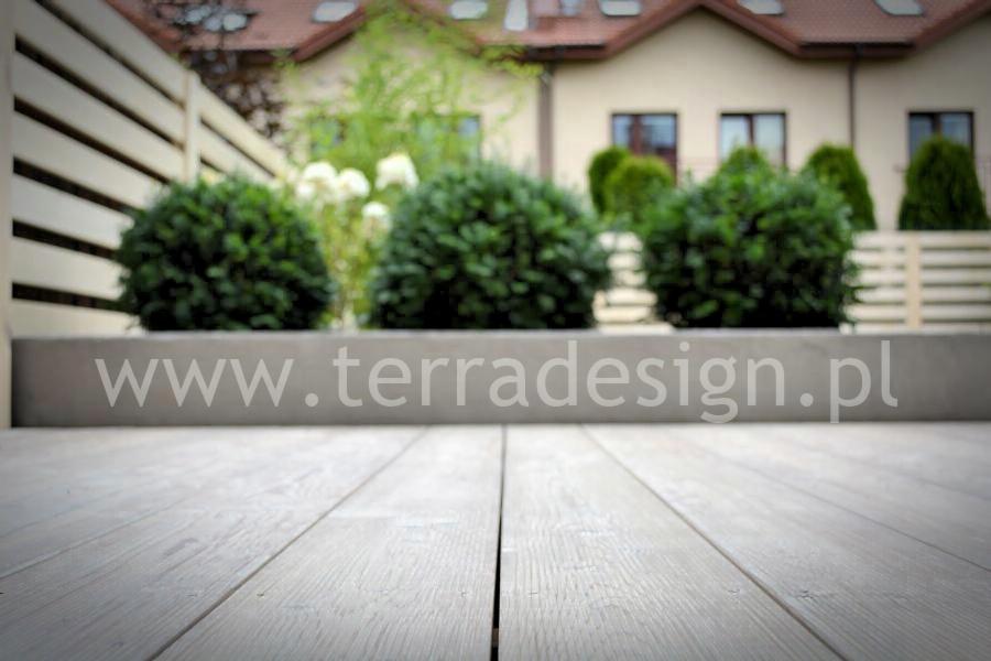 Jasnoszary taras z desek kompozytowych, z widoczną nazwą firmy, w tle kuliste krzewy i jasny płot lamelowy.
