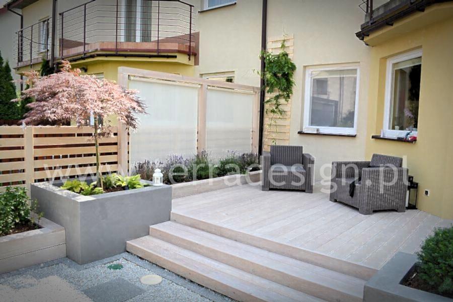 Jasny taras kompozytowy z trzema stopniami, dwoma szarymi fotelami z technorattanu, betonową donicą z drzewkiem i niską drewnianą ażurową zabudową w tle.