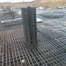 Zbrojenie fundamentu z pionowymi prętami zbrojeniowymi, z widoczną siatką zbrojeniową i elementami szalunkowymi w tle.