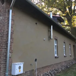 Odnowiona elewacja budynku z nowymi oknami, wykończona tynkiem w kolorze beżowym, z widocznym kominem i rynną oraz fragmentem ceglanej ściany szczytowej.