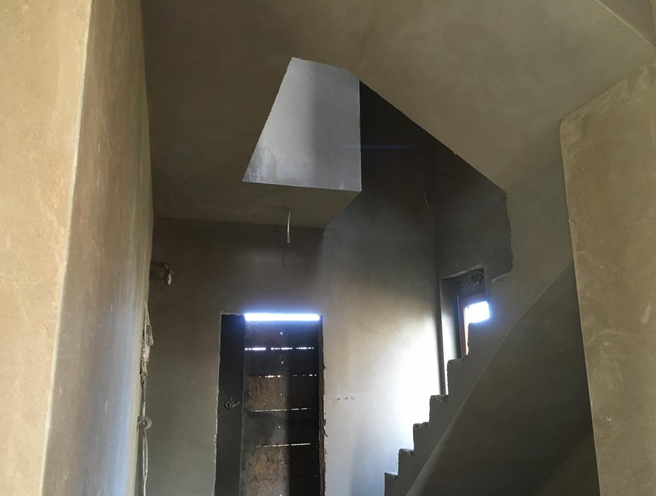 Wnętrze domu w trakcie prac wykończeniowych, widoczne świeżo otynkowane ściany i schody, z fragmentami surowego drewna w tle, oświetlone wpadającym światłem słonecznym.