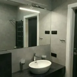 Nowoczesna łazienka z szarymi płytkami, białym umywalką nablatową, dużym lustrem z oświetleniem LED i czarnym grzejnikiem drabinkowym.