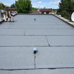 Widok z góry na płaski dach pokryty szarą papą, z widocznymi kominami, anteną satelitarną i elementami obróbki blacharskiej. Tło stanowią budynki i zieleń.