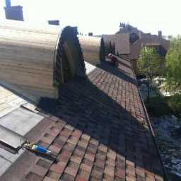 Wymiana dachu z drewnianą konstrukcją łukową i układaniem gontów bitumicznych, widoczny pracownik w trakcie pracy oraz narzędzia dekarskie na powierzchni dachu.