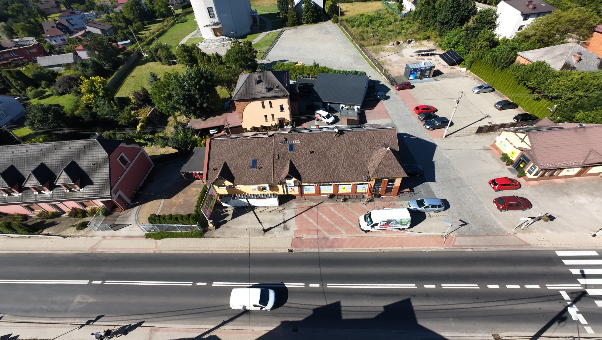 Widok z góry na budynek z brązowym dachem, otoczony zielenią i ulicą z samochodami. Dach po renowacji, widać detale wykończenia i otoczenie budynku.