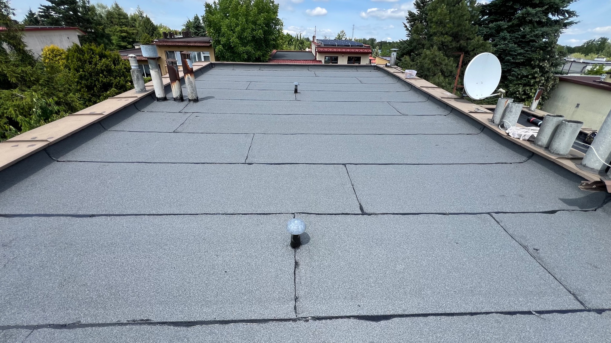 Widok z góry na płaski dach pokryty szarą papą, z widocznymi kominami, anteną satelitarną i elementami obróbki blacharskiej. Tło stanowią budynki i zieleń.