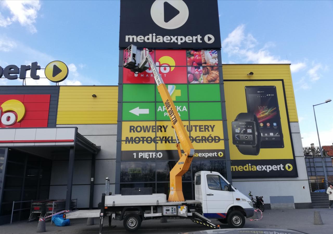 Żółty podnośnik koszowy na białej ciężarówce z podnośnikiem teleskopowym na tle ściany budynku Media Expert z reklamami i logotypami.