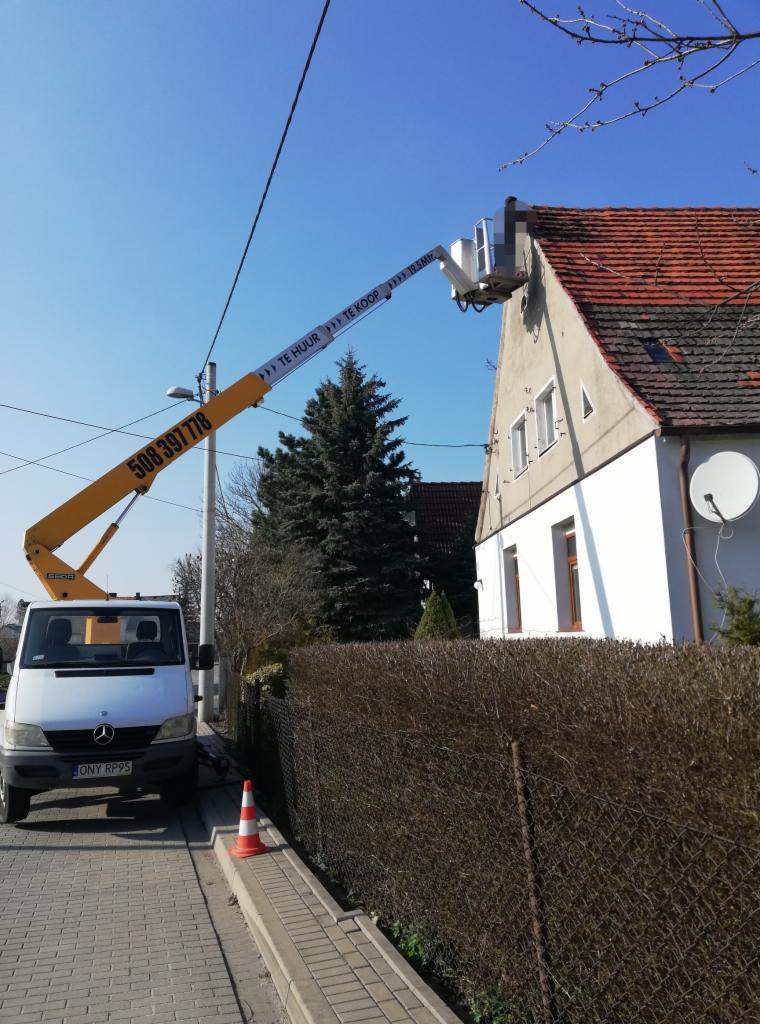 Żółty podnośnik koszowy z numerem telefonu i napisem 'TE HUUR TE KOOP' ustawiony przy białym domu z czerwoną dachówką, na tle błękitnego nieba.