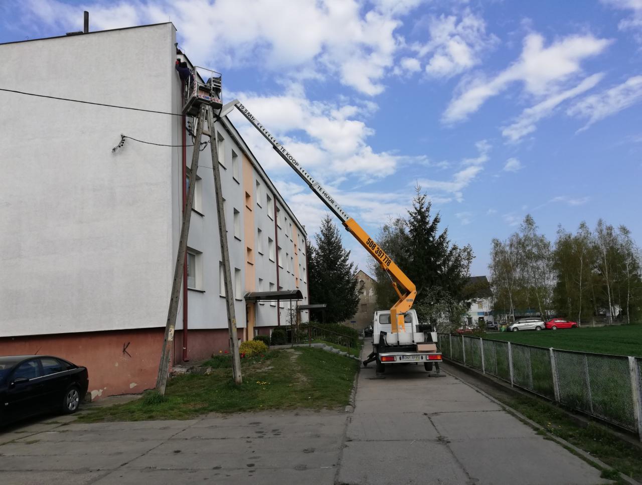 Żółty podnośnik koszowy na pojeździe, ustawiony przy ocieplonym bloku mieszkalnym, z pracownikiem w koszu na wysokości elewacji.