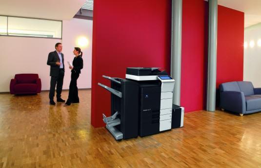 Nowoczesna, czarno-biała kserokopiarka A3/A0 marki Konica Minolta w biurze z parkietem, czerwonymi ścianami i rozmawiającymi osobami w tle.