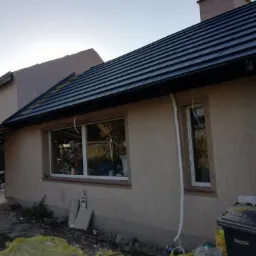 Nowy dach z czarnej dachówki ceramicznej na domu jednorodzinnym, widoczne rynny i elewacja w trakcie wykończenia, teren wokół domu częściowo zabezpieczony folią.