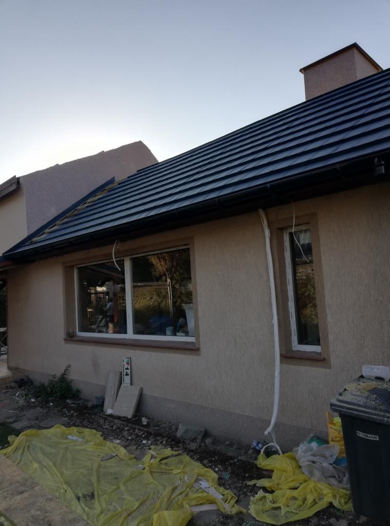 Nowy dach z czarnej dachówki ceramicznej na domu jednorodzinnym, widoczne rynny i elewacja w trakcie wykończenia, teren wokół domu częściowo zabezpieczony folią.