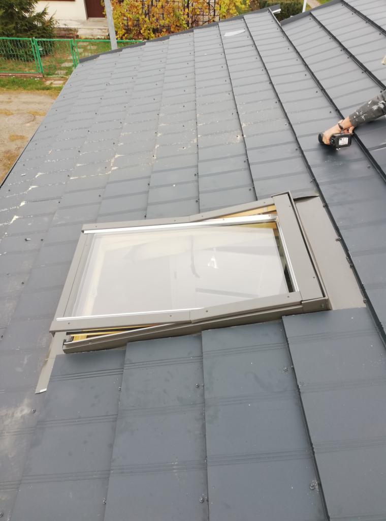 Montaż okna dachowego na grafitowym dachu z blachodachówki modułowej, widoczne narzędzie w dłoni dekarza, jesienne drzewa w tle.