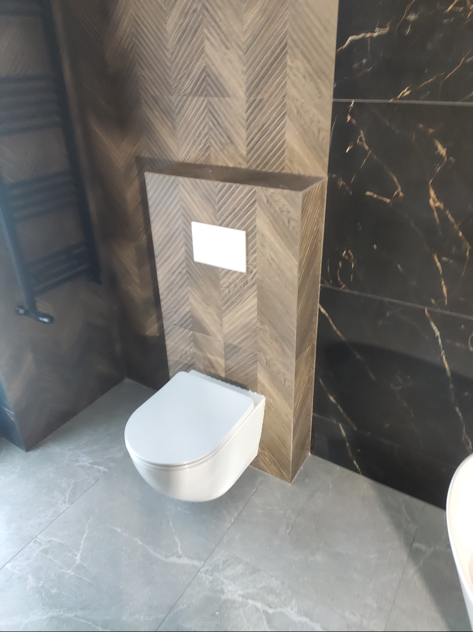 Nowoczesna toaleta podwieszana z zabudową w płytkach imitujących drewno ułożonych w jodełkę, obok ściana z czarnych płytek z marmurkowym wzorem. Szara podłoga.