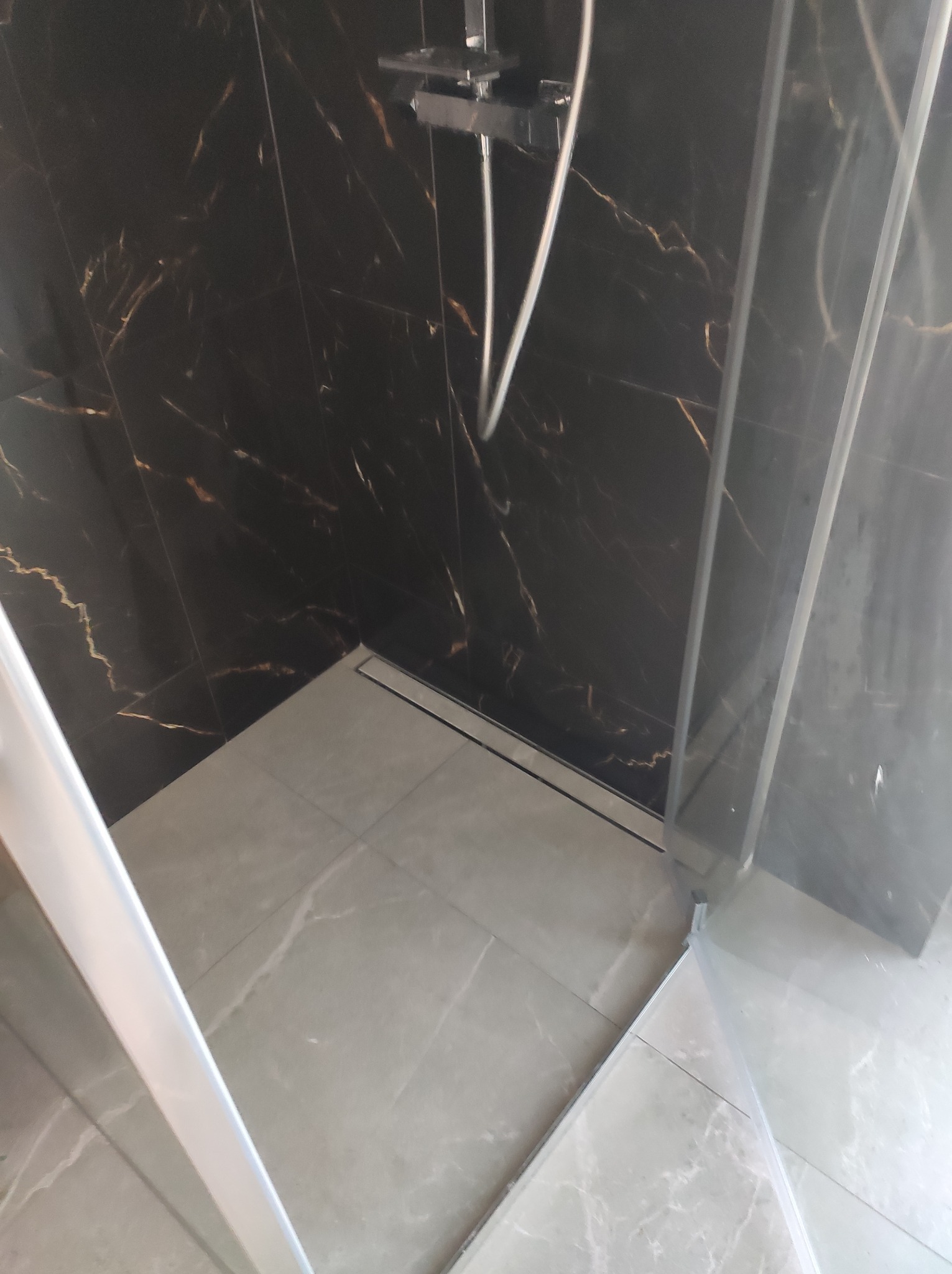 Nowoczesna kabina prysznicowa z czarnymi płytkami imitującymi marmur i szarymi płytkami podłogowymi z odpływem liniowym. Minimalistyczny design.