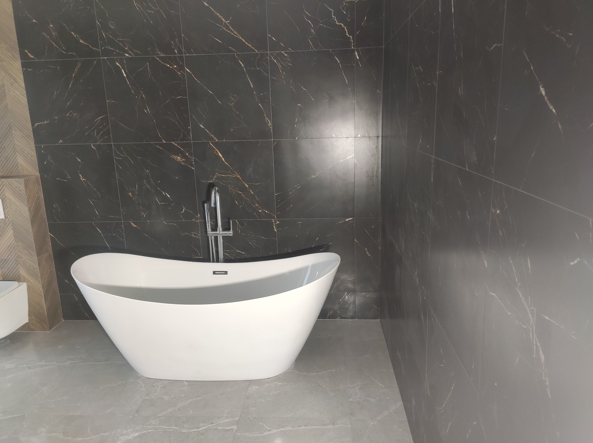 Biała, wolnostojąca wanna w łazience z ciemnymi płytkami imitującymi marmur i szarą podłogą. Nowoczesny design i chromowana bateria.